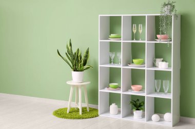 Mutfaktaki yeşil duvarın yanında çanak çömlek ve bitki bulunan modern raf ünitesi.