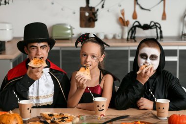 Cadılar Bayramı için giyinmiş çocuklar mutfakta pizza yiyorlar.