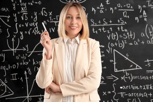 Sınıfta tahtanın yanında tebeşirle tebeşir tutan olgun kadın matematik öğretmeni.