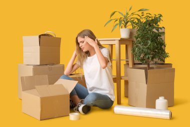 Stresli genç kadın eşyalarını sarı arka plandaki karton kutulara dolduruyor. Taşınma konsepti