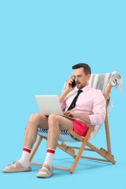Dizüstü bilgisayarlı komik bir iş adamı güvertede oturuyor ve mavi arka planda cep telefonuyla konuşuyor.