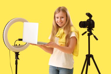 Genç blogcu elinde halka ışığı ve sarı arka planda fotoğraf makinesi olan dizüstü bilgisayar tutuyor