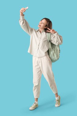 Sırt çantalı mutlu kız öğrenci zafer işareti yapıyor ve mavi arka planda cep telefonuyla selfie çekiyor.