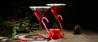Lezzetli kırmızı margarita kadehleri, hediye, baston şekerler ve koyu arkaplanda Noel ağacı dalları.