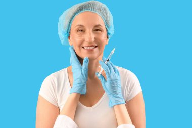 Olgun kadın ve güzellik uzmanının ellerinde mavi arka planda dolgu iğnesi, yakın plan.