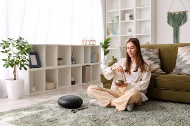 Young woman with Tibetan singing bowl sitting on carpet at home clipart