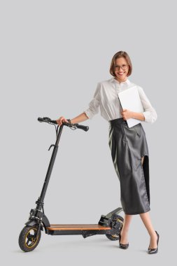 Beyaz arka planda dizüstü bilgisayarı ve elektrikli scooterı olan mutlu genç bir iş kadını.