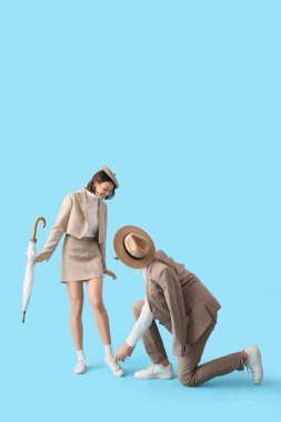 Genç adam mavi arka planda kız arkadaşının ayakkabı bağcıklarını şemsiyeyle bağlıyor.