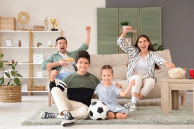 Futbol topu ve atıştırmalıklarla mutlu bir aile evde futbol maçı izliyor.