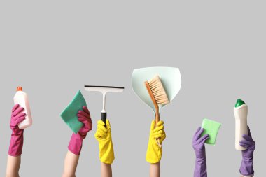 Deterjan, paçavra, faraş, süpürge ve gri arka planda cam sileceği olan lastik eldivenli kadın elleri.