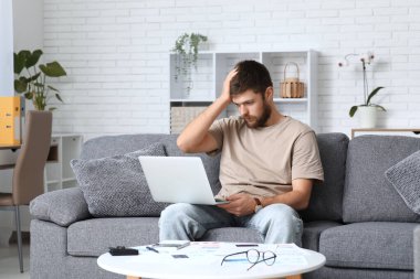 Evdeki kanepede dizüstü bilgisayar kullanarak borç batağındaki stresli genç adam