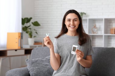 Glikoz testi bantları ve glukometresi olan şeker hastası genç bir kadın evdeki kanepede oturuyor.