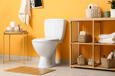 Tuvaletin içinde beyaz klozet, sehpa ve turuncu duvarın yanında raf ünitesi var.