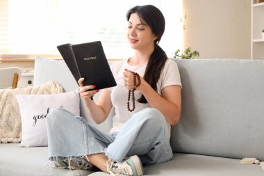 Evdeki kanepede kutsal kitap okuyan dindar genç bir kadın.