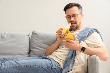 Cep telefonlu genç adam evdeki kanepede muz yiyor.