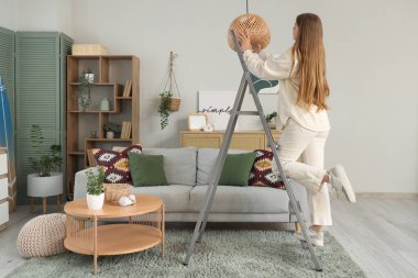 Genç bir kadın tavan lambasına ampul takıyor. Evinde, arka planda.