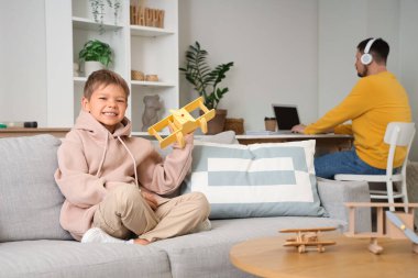 Küçük çocuk oyuncak oynuyor ve babası kulaklıkla evde dizüstü bilgisayarla çalışıyor.