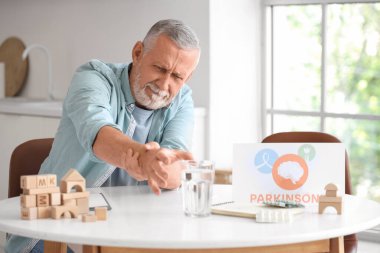 Senior man suffering from Parkinson syndrome at table in kitchen clipart