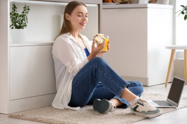 Genç, mutlu bir kadın dizüstü bilgisayar, bir bardak meyve suyu ve lezzetli bir tost ve evde taze tereyağı tutuyor.