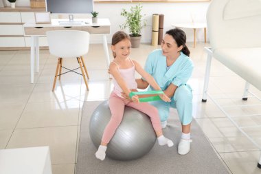 Kadın osteopat ve elastik bantlı küçük bir hasta klinikte topa uygun egzersiz yapıyorlar.