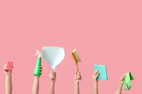 Руки женщины держат бутылку моющего средства, тряпку, резиновые перчатки, совок, метлу и губку на розовом фоне