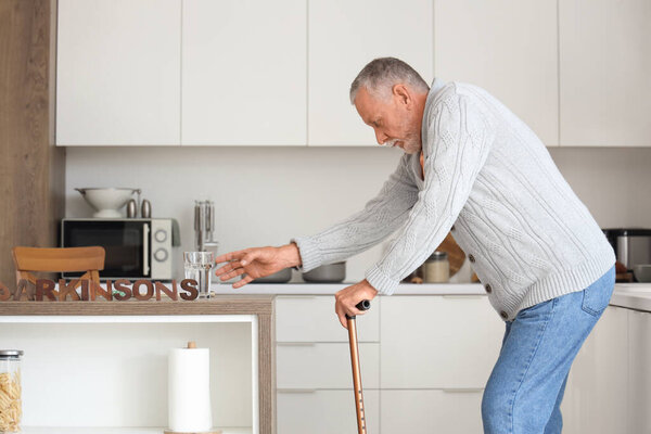 Senior man suffering from Parkinson syndrome taking glass of water from table in kitchen