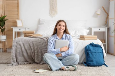 Sırt çantası, kitapları ve dizüstü bilgisayarı olan mutlu bir kız öğrenci evde oturuyor.