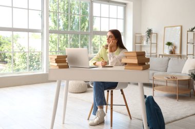 Kitap, defter ve dizüstü bilgisayarla evde ders çalışan yorgun bir kız öğrenci.