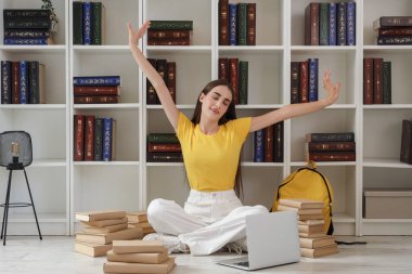 Sırt çantası, kitap ve dizüstü bilgisayarı olan mutlu bir kız öğrenci kütüphanede rafların yanında ders çalışıyor.