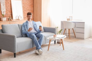 Evdeki gri koltukta kahve fincanı kullanan genç adam.