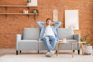 Evdeki gri koltukta oturan genç adam.