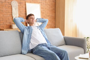 Evdeki gri koltukta oturan genç adam.