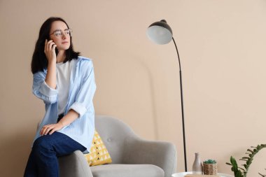 Evdeki bej duvarın yanındaki koltukta cep telefonuyla konuşan genç bir kadın.