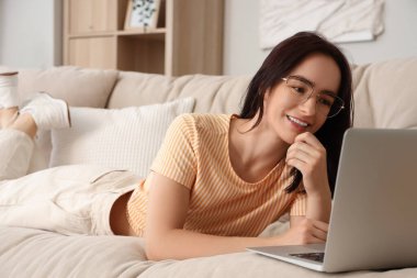 Evdeki bej koltukta dizüstü bilgisayar kullanan genç bir kadın.