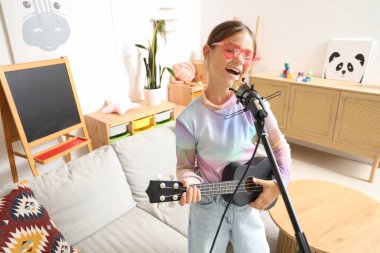 Evde mikrofonlu küçük bir kız gitar çalıyor.
