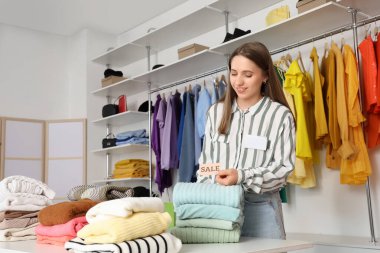 Butikte katlanmış kıyafetlerin yanında satış tabelası olan kadın satıcı.