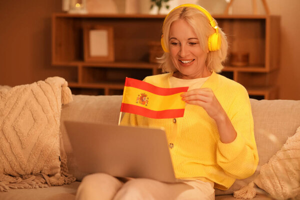Mature woman with headphones and flag of Spain learning language online at home in evening