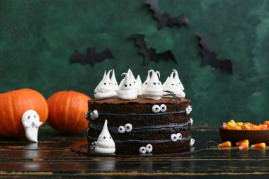 Tatlı Cadılar Bayramı pastası, yeşil arka planda siyah ahşap masada hayaletler, balkabakları ve bir kase lezzetli şekerlerle süslenmiş.