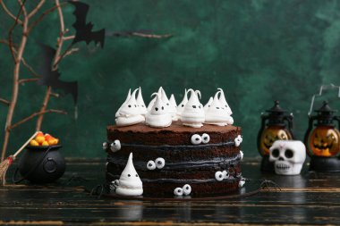 Tatlı cadılar bayramı pastası, yeşil arka planda siyah ahşap masada hayaletler, şekerler, örümcekler ve fenerlerle süslenmiş.