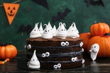 Tatlı Cadılar Bayramı pastası, yeşil arka planda siyah ahşap masada hayaletler ve balkabaklarıyla süslenmiş.