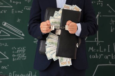 Çantayla kelepçelenmiş erkek öğretmen ve kara tahtanın yanında rüşvet, yakın plan. Yolsuzluk kavramı