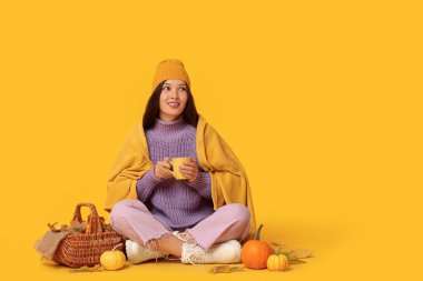 Bir fincan kahve, hasır sepet, sonbahar yaprakları ve sarı arka planda duran taze balkabaklarıyla güzel bir Asyalı kadın.