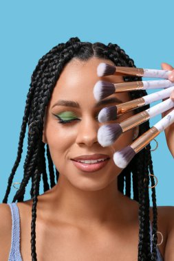 Saçları örgülü, makyajlı, mavi arka planda fırça tutan genç bir Afro-Amerikalı kadın.