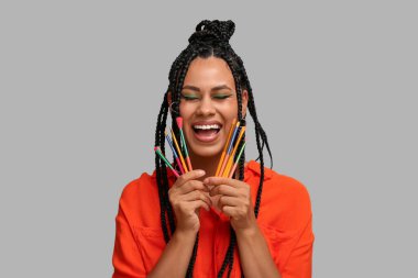 Saçları örgülü ve güzel makyajlı, gri arka planda fırçaları olan mutlu, Afro-Amerikalı bir kadın.