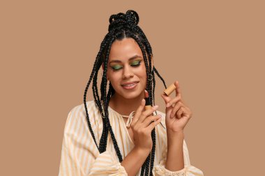Saçları örgülü ve makyajlı genç bir Afro-Amerikalı kadın kahverengi arka planda ruj tutuyor.