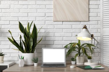 Ofiste beyaz tuğla duvarın yanında boş dizüstü bilgisayarı, lambası ve yeşil bitkileri olan işyeri