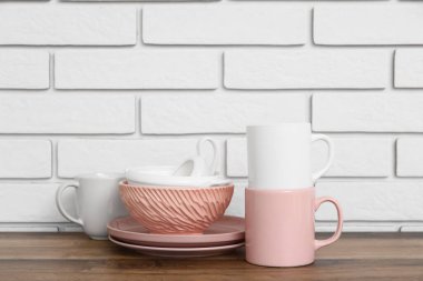 Bardaklar, tabaklar ve kaseler beyaz tuğla duvarın yanındaki ahşap masada.