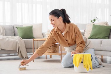 Genç bir kadın evde fırçayla halı temizliyor.