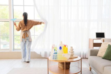 Evde perdeleri açık, arka manzaralı genç bir kadın.