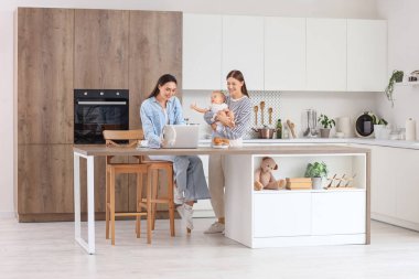 Küçük bebekleriyle mutfakta dizüstü bilgisayar kullanan genç lezbiyen çift.